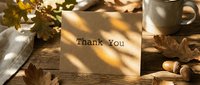 Thank you card on rustic wooden table with autumn leaves, acorns, and coffee mug in warm sunlight, cozy fall seasonal greeting concept.