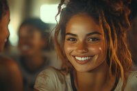 Young African American woman with dreadlocks and natural makeup smiling warmly at camera, illuminated by soft golden light in casual social setting.