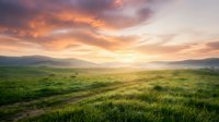 Scenic rolling grassland valley at sunrise with golden light breaking through dramatic clouds over distant mountains and lush green meadow.