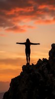 Woman silhouette with arms outstretched standing on rocky cliff against dramatic orange sunset sky expressing freedom and achievement.