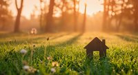 Small wooden house model on green grass field during golden hour sunset with warm sunlight and bokeh background for real estate concepts.