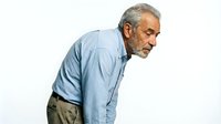 Senior man with gray beard and hair, wearing light blue button-up shirt and olive pants, leaning forward with hands on knees in contemplative pose against white background.