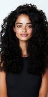 Young mixed race woman with voluminous curly black hair and natural makeup wearing black top against light background, looking directly at camera with confident smile.