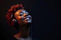 Young African American woman with red curly hair wearing headphones, smiling joyfully while listening to music against dark background with colorful lighting.