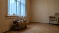 Vintage leather suitcase sits on wooden floor in empty room with beige walls, vertical blinds at window, and upholstered chair. Relocation, moving day, travel concept.