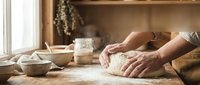 Hands kneading fresh dough on wooden table in rustic kitchen with flour, bowls and natural lighting for homemade bread baking and cooking preparation.