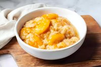 Fresh peach cobbler served in white ceramic bowl on wooden board, topped with golden crust and juicy fruit slices in sweet syrup. Homestyle comfort dessert.