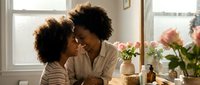 Adult woman of African descent with curly dark hair embracing young child with similar hair texture in bright, sunlit room with flowers and windows, depicting tender family bonding moment.