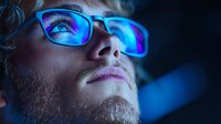 Young bearded man wearing glasses illuminated by blue light from screen, close-up portrait with neon glow reflection showing digital lifestyle at night.