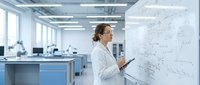 Female scientist in white lab coat analyzing data on whiteboard in modern research laboratory with equipment and bright lighting.