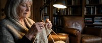 Senior woman with gray hair and light skin tone knitting with needles and yarn in cozy home library setting with warm lighting and leather furniture.
