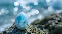Serene larimar gemstone resting on coastal rock with sparkling ocean bokeh in background, showcasing natural blue mineral specimen against blurred aqua seascape.