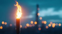 Burning torch flame against blurred city lights at dusk creates atmospheric mood with bokeh effect and dramatic orange glow against teal blue sky.
