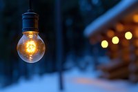 Glowing light bulb illuminated against snowy winter evening background with warm bokeh lights creating cozy atmospheric scene. Outdoor holiday decoration.