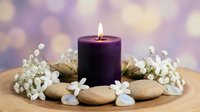 Purple candle burning surrounded by white flowers and smooth stones on wooden surface with soft bokeh background for spa relaxation and wellness.