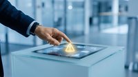 Businessman hand touching glowing light bulb hologram on digital tablet with blurred office skyscraper background, representing innovation and technology solutions.