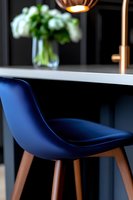 Modern navy blue bar stool with wooden legs against dark counter, copper pendant lamp above, and blurred white flowers in background creating luxe ambiance.