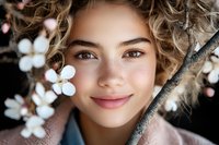 Young woman with natural curly hair and gentle smile surrounded by white spring blossoms, wearing pink coat, close up portrait with soft natural lighting.