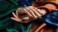 Elegant female hands with dark manicure and gold rings resting on colorful satin fabric in teal, rust and blue tones.