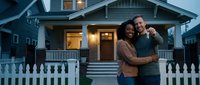 Happy interracial couple embracing in front of their new home at twilight with warm porch lights creating welcoming atmosphere for real estate and homeownership concepts.