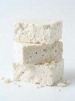 Three stacked blocks of fresh feta cheese with crumbly texture and scattered crumbs on white background, suitable for food photography and culinary design projects.