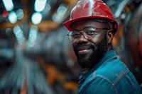 Smiling African American male industrial worker wearing red hard hat, safety glasses and work uniform in manufacturing facility with bokeh background.