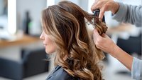 Professional hairstylist styling woman's wavy brown hair with blonde highlights in modern salon, creating elegant curls for special occasion.