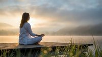 Young woman practicing yoga meditation on wooden dock at sunrise over misty lake with mountains in background creating peaceful wellness atmosphere.