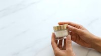 Hands holding luxury cream jar with gold lid against marble background for skincare and cosmetics marketing.