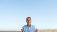 Middle-aged man with gray hair and light skin tone wearing light blue shirt gestures welcomingly against clear sky and agricultural field backdrop.
