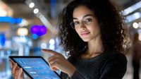 Young mixed race businesswoman with curly hair and glasses analyzing financial data on digital tablet with glowing charts against blurred office background at night.
