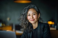 Confident Asian businesswoman with gray curly hair smiling in dimly lit cafe while working on laptop, portraying professional success and modern leadership.