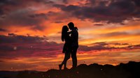 Silhouette of couple kissing against dramatic orange and red sunset sky with clouds, romantic outdoor scene for love and relationship concepts.