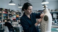 Female fashion designer working on dress form in modern studio workspace with industrial lighting and fabric shelves in background.