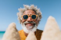 Senior African American man with gray curly hair and beard wearing yellow sunglasses smiles joyfully at beach on sunny summer day against blue sky and ocean background.