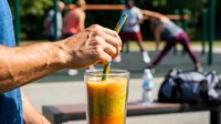 Man holding straw in glass of cold beverage at outdoor sports court with athletes exercising in background, refreshment during fitness activity.