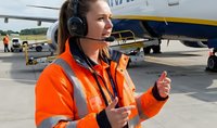 Female airport ground crew worker in orange safety vest with headset giving thumbs up on tarmac with aircraft in background for aviation industry concepts.