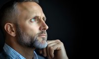 Profile of contemplative mature Caucasian man with gray beard wearing blue checkered shirt against dark background, hand on chin in thoughtful pose looking upward.