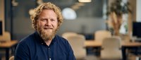 Smiling bearded middle-aged Caucasian businessman with curly hair in modern office workspace for corporate headshot and professional marketing materials.