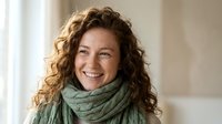Happy middle-aged woman with curly brown hair wearing green knit scarf smiling warmly at camera in bright indoor setting for lifestyle content.