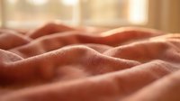 Soft coral pink blanket with gentle folds creating cozy texture in warm sunlight streaming through window for home comfort and relaxation concepts.