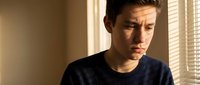Young Caucasian man in dark blue shirt looking down with concerned expression in warm sunlit room with window blinds creating dramatic lighting shadows.