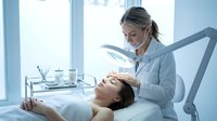 Professional dermatologist examining young woman's face with magnifying lamp in modern clinic for skincare treatment and cosmetic consultation.