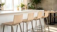 Modern bar stools with wooden seats line a minimalist kitchen counter near bright windows, featuring contemporary Scandinavian design for residential or commercial interior spaces.