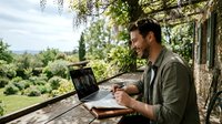 Man working on laptop outdoors in garden setting, taking notes in notebook while sitting at wooden table near stone house with green landscape background.