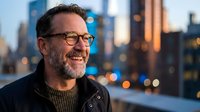 Mature Caucasian businessman with glasses and beard smiling outdoors at dusk with city skyline background. Professional portrait for corporate marketing.