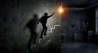 Businessman climbing concrete stairs toward glowing light bulb representing success, achievement and career growth in modern industrial interior.