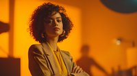 Young African American businesswoman with curly hair standing confidently with arms crossed against vibrant orange wall with dramatic shadows and lighting.