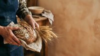 Farmer holding fresh baked bread loaf and wheat stalks against rustic background. Suitable for bakery, agriculture, and food industry marketing materials.