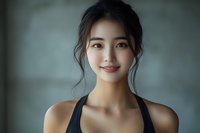 Young Asian woman with gentle smile wearing black athletic top, natural makeup and hair in casual updo against soft blurred background. Wellness and fitness concept.